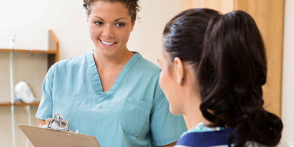 A woman wearing scrubs and carrying a clipboard speaks with another woman